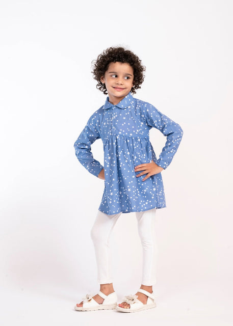 A young child with curly dark hair stands against a plain white studio background wearing a blue long-sleeve collared shirt with a white star pattern, white leggings, and white sandals.